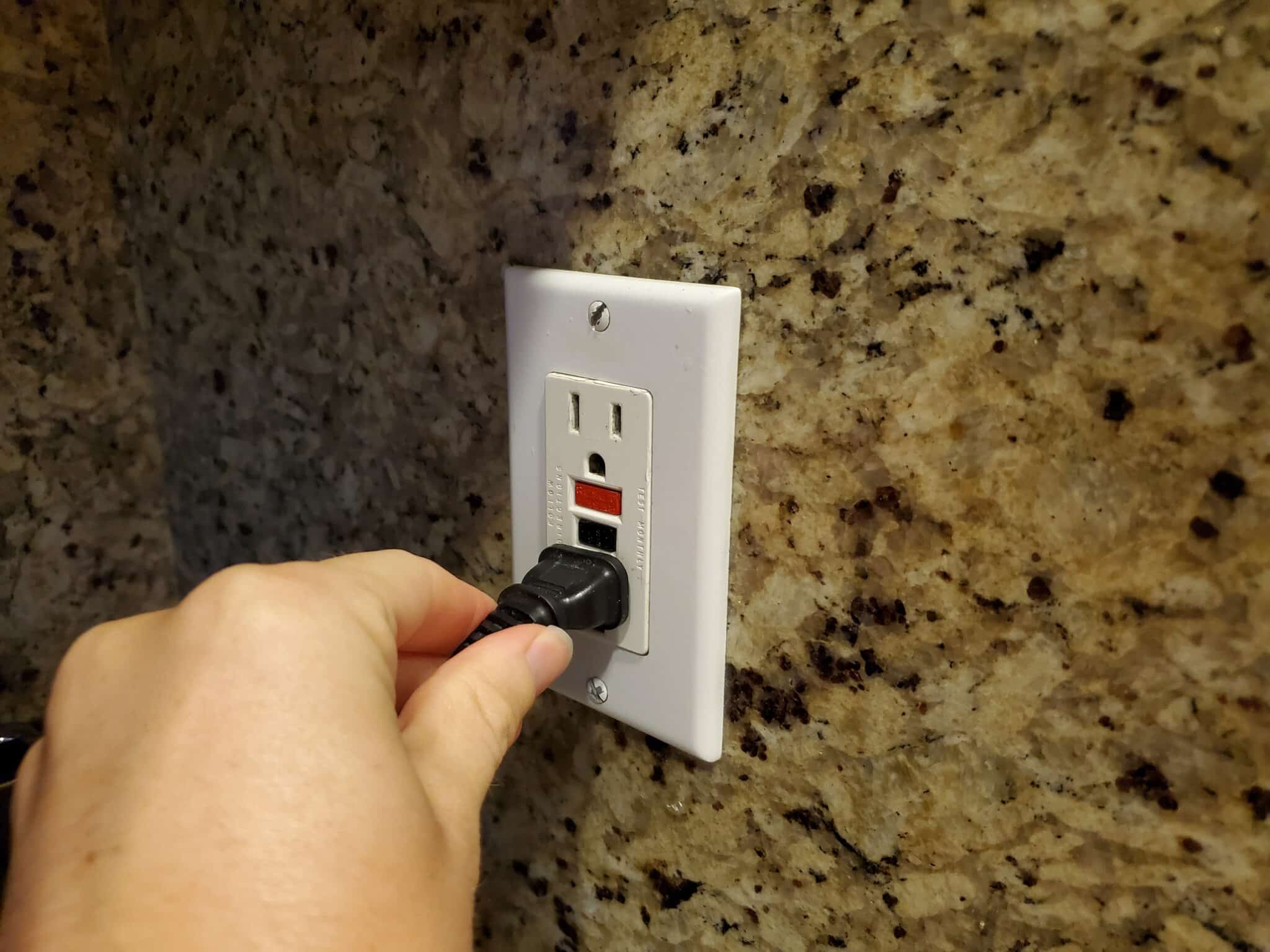 A personal perspective image of a man's hand plugging an electrical cord into a Ground Fault Circuit Interrupter (GFCI) electrical outlet on the wall.