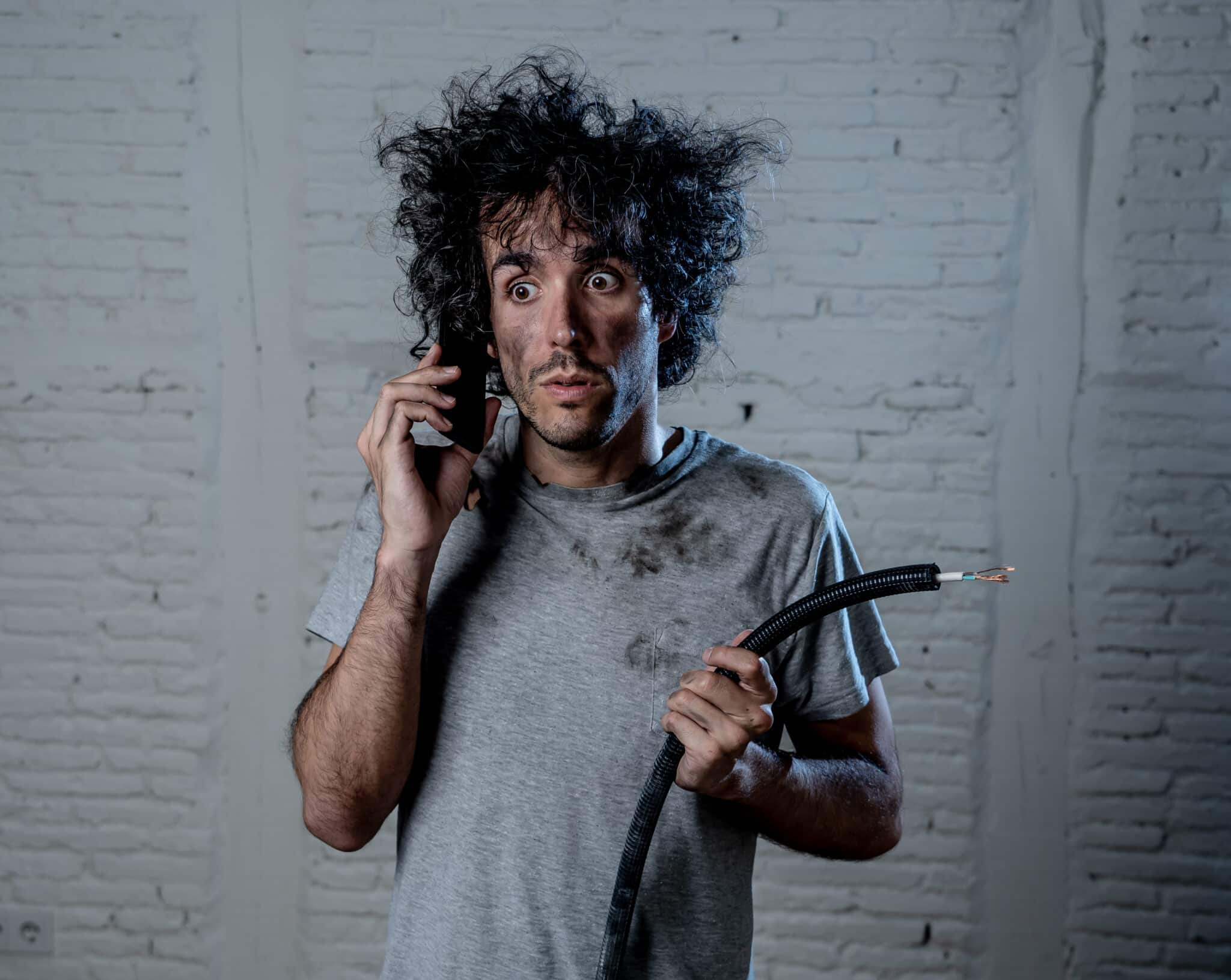 A portrait of a young man holding an electrical cable after a domestic accident, with a dirty, burnt, and funny expression on his face. He's calling desperately with a mobile phone, asking for help. This scene highlights electrical emergencies, illustrating the need for prompt assistance in such situations.