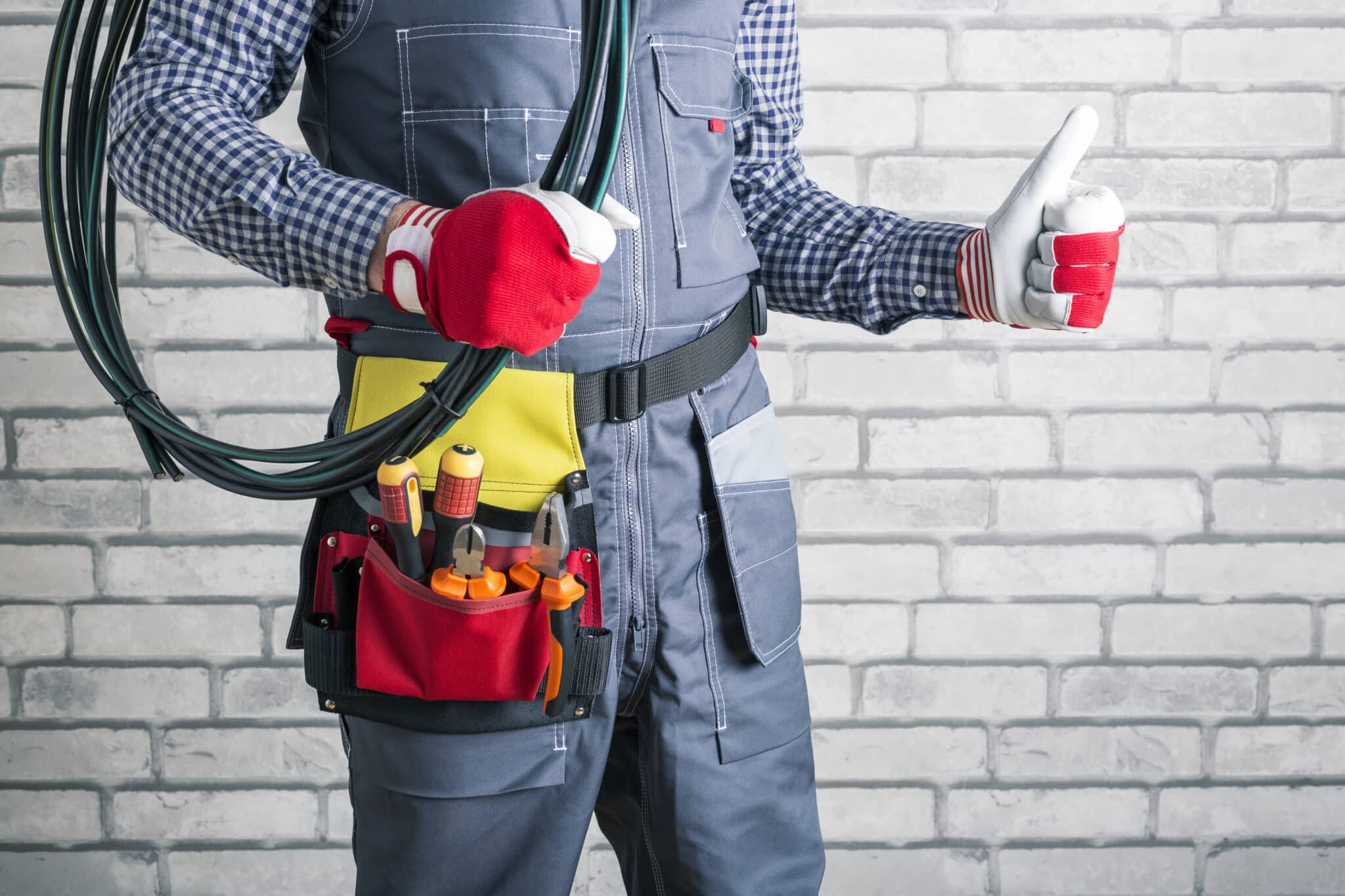An electrician in uniform, holding rolled wires and giving a thumbs-up against a brick wall background.